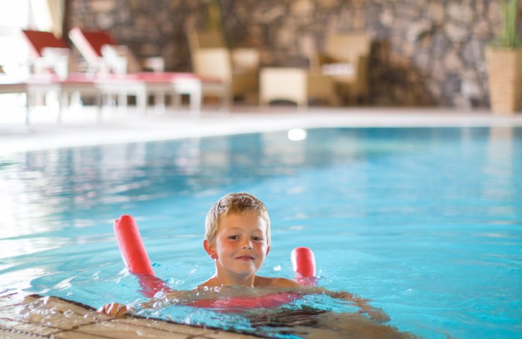 Boy swimming with a pool noodle in the hotel's indoor pool, loungers by a stone wall behind